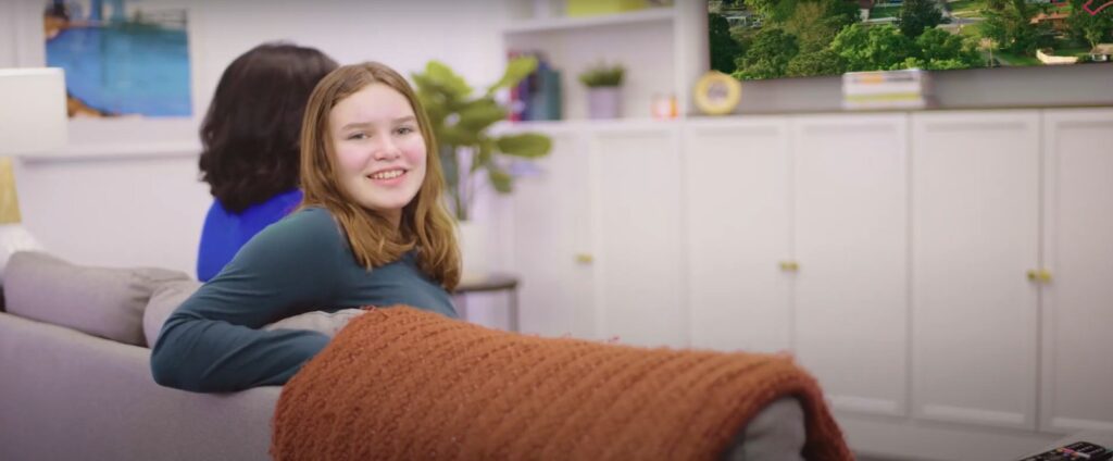 Image of a mother and daughter watching TV. Daughter has leaned over the couch to smile at the camera.